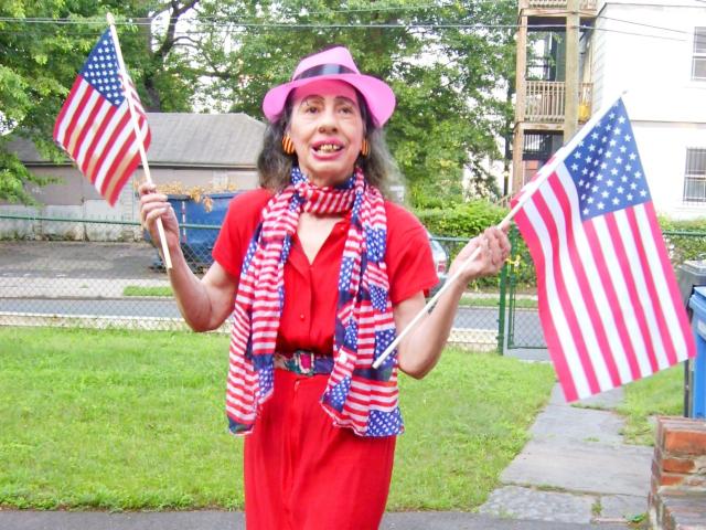 ElenahHottie vêtue de rouge, avec un chapeau rose, une écharpe rayée, tenant deux drapeaux américains, souriante, debout sur le trottoir, avec de l'herbe et une maison en briques en arrière-plan, ayant l'air joyeuse..