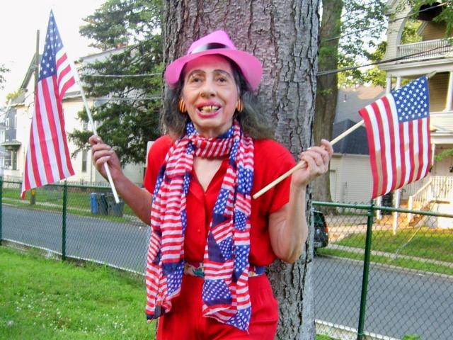 ElenahHottie com chapéu rosa, vestido vermelho, cachecol listrado, segurando duas bandeiras americanas, em pé perto de uma árvore, grama e cerca, sorrindo com um sorriso largo, durante o dia, em um ambiente suburbano..