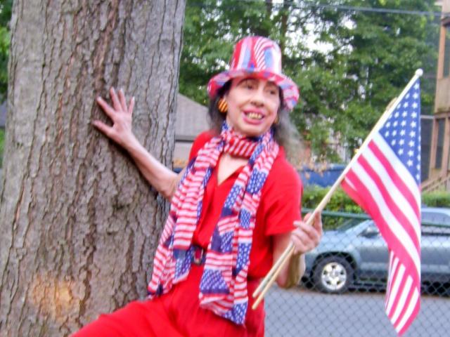 ElenahHottie vestindo um traje vermelho, um chapéu com a bandeira americana e um cachecol listrado, segurando uma bandeira, apoiada em uma árvore, sorrindo, com cabelo grisalho e batom vermelho..