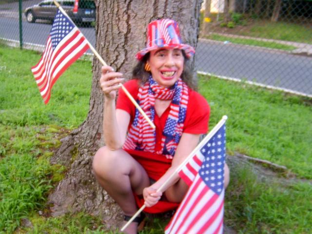 ElenahHottie, seduta a squat vicino a un albero, indossa un vestito rosso, una sciarpa a righe e un cappello, tiene due bandiere americane, sorride con i capelli castani ricci, rossetto rosso e glutei visibili..