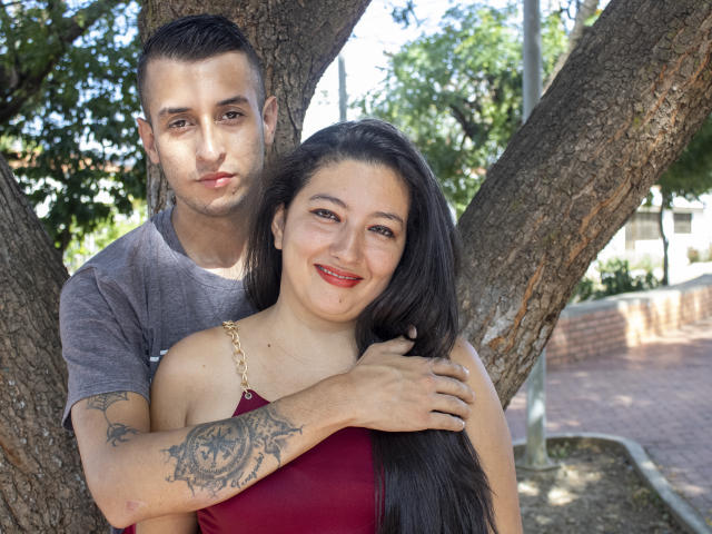 MaxAndViolet com cabelo preto longo e vestido vermelho, tatuagem no braço direito, sorrindo, em pé em frente a uma árvore. MaxAndViolet com cabelo castanho curto, camisa cinza, braços ao redor dela..