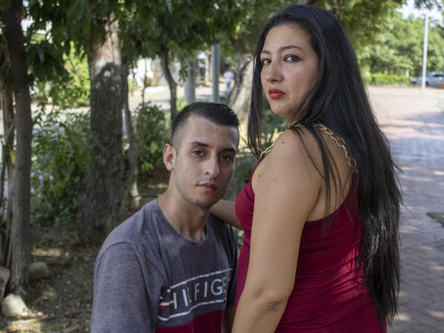 MaxAndViolet, con lunghi capelli neri e un vestito rosso, è in piedi accanto a MaxAndViolet, con capelli neri corti e una camicia grigia, che appare seria. Entrambe hanno la pelle media e rossetto rosso..
