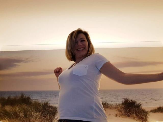 OscarBella, con un corte de pelo rubio, una camisa blanca y senos pequeños, sonriendo, con los brazos levantados, de pie frente a una foto del atardecer..