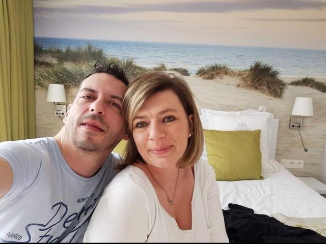 OscarBella, com cabelo castanho curto, vestindo branco, e um homem com cabelo cacheado escuro, vestindo uma camisa azul clara, ambos sorrindo em uma cama com travesseiros verdes..