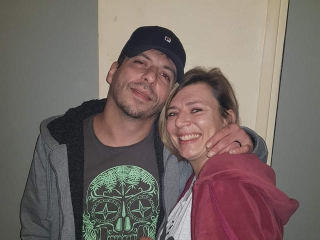OscarBella, een man met een zwarte pet en grijze hoodie, staat met zijn arm om OscarBella, een lachende vrouw in een rode hoodie en grijze shirt..