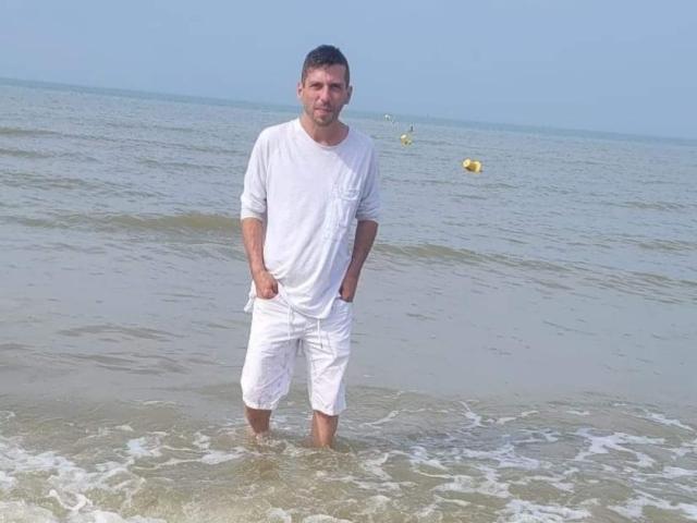 OscarBella está de pie en aguas poco profundas, vistiendo una camisa blanca y pantalones cortos mojados, con las manos en los bolsillos, y un mar tranquilo y boyas amarillas al fondo..
