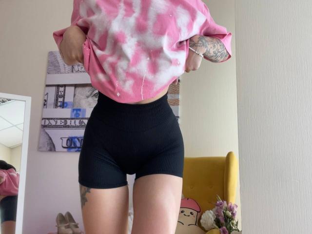 Tsukerberg in pink tie-dye shirt, black ribbed shorts, tattooed arms, lifting shirt, showing midriff, yellow chair with pink Hello Kitty pillow in background.