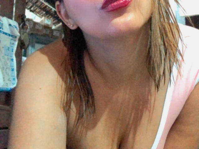 Sarita Velez, con el cabello castaño mojado, lápiz labial rojo y una blusa rosa y blanca, que muestra su escote y está ligeramente abierta. Se ven los senos, mirando hacia abajo con una expresión juguetona y ligeramente fruncida..