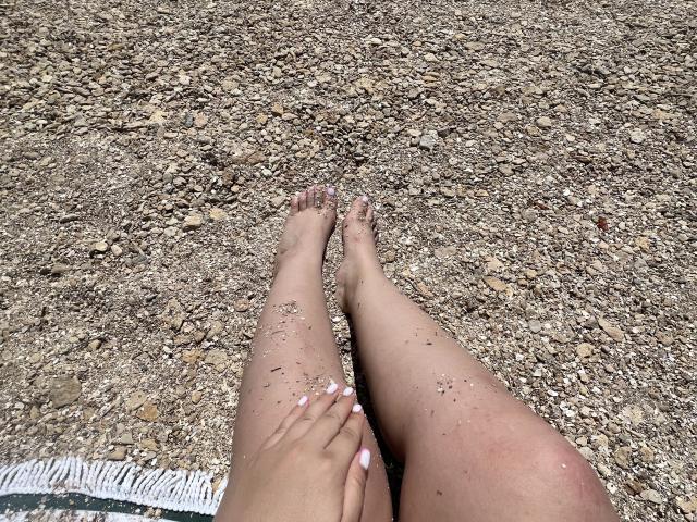 Barbarica's sand-covered legs and feet are in the foreground, showing pink nail polish and small pebbles stuck to her skin. Her hands rest gently on her thighs.