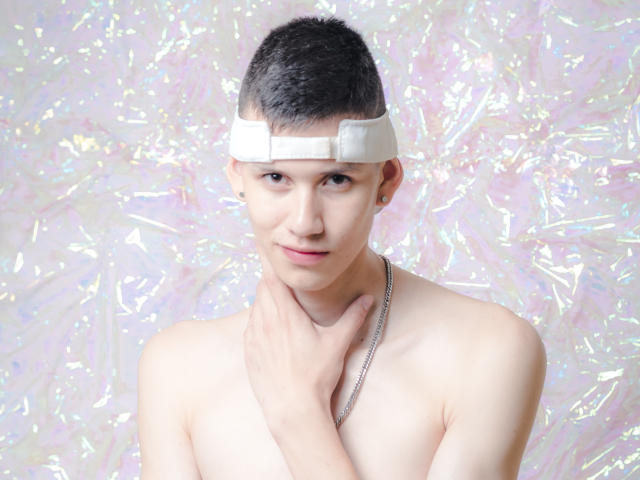 JeanKarloLeone with short black hair, wearing a white headband, shirtless, touching his chin, silver necklace, and a small earring, against a shiny, iridescent background.