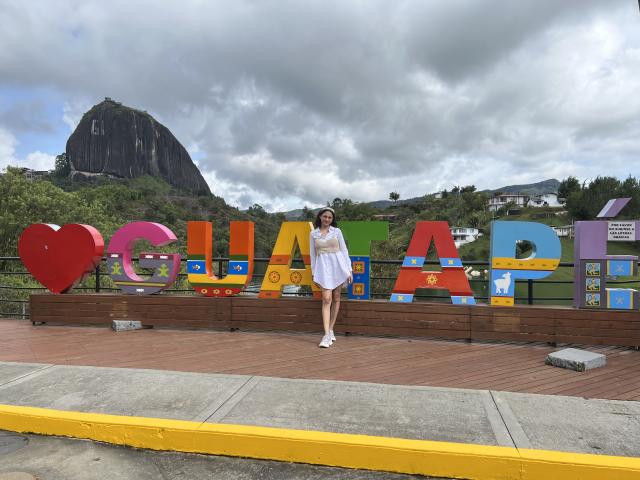 EstefaniaRamosHot stands in front of colorful letters with a heart, wearing a white dress and white shoes, under a cloudy sky, with a tall mountain in the background.