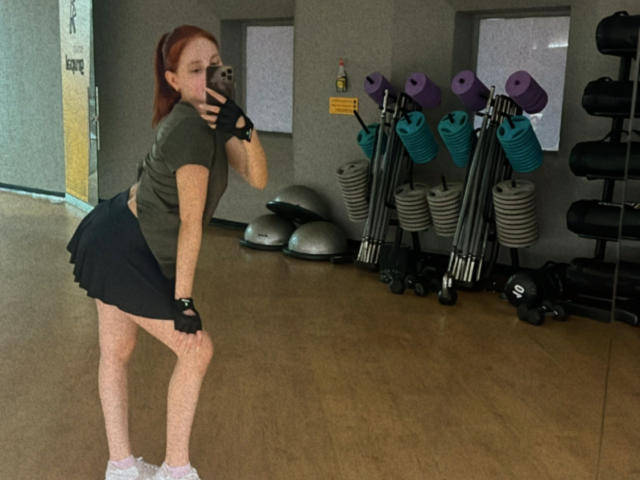 EstefaniaRamos, attraente, con capelli rossi, maglietta verde, gonna nera, calze bianche, che tiene un telefono, si piega e mostra il sedere, in palestra con manubri e palle da ginnastica..