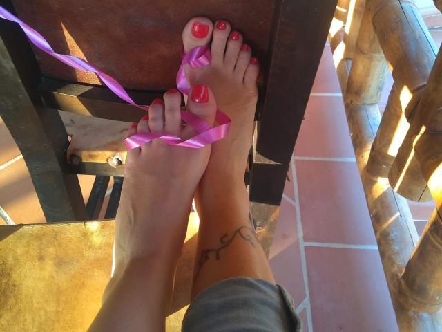 I piedi di Karol Meyer, con le unghie dei piedi dipinte di rosso, un nastro rosa legato all'alluce e un piccolo tatuaggio sulla caviglia, in piedi su un portico piastrellato..