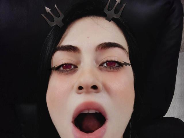 Il volto di KarolMeyer, con capelli neri, occhi rossi e eyeliner nero, indossa una corona nera, la bocca è aperta, con un'espressione sorpresa o eccitata, su uno sfondo scuro..