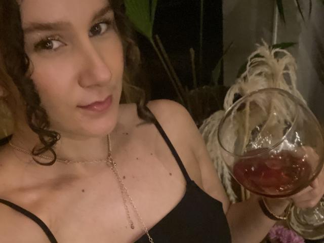 Aldhara, con i capelli castano ricci, tiene in mano un bicchiere di vino rosso, indossa una maglia nera e una delicata collana, mostrando il seno e un leggero sorriso..