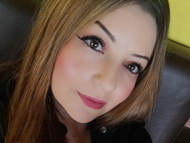 Il viso di NatalyHami, con i capelli castani, l'eyeliner scuro, i grandi occhi marroni, il rossetto rosa e un leggero sorriso, indossa una camicia nera e siede su una poltrona in pelle marrone..