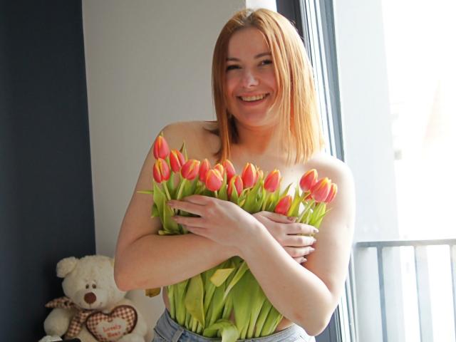 LaylaXDream with red hair, smiling, holding a bouquet of orange and yellow tulips, arms crossed, topless, wearing blue jeans, standing by a window, white teddy bear in background.