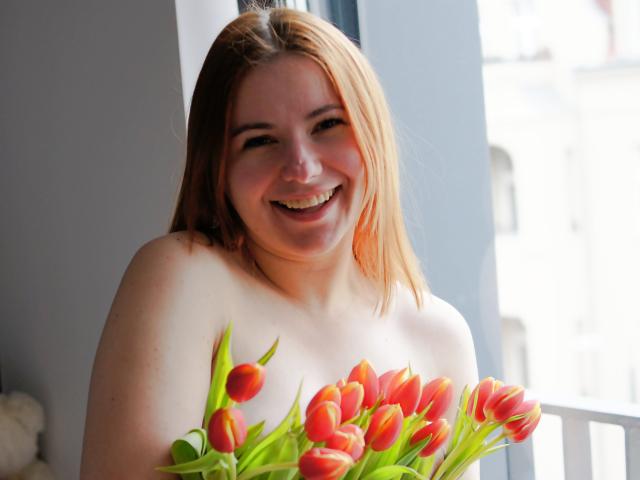 LaylaXDream, con le spalle scoperte, sorridente, che tiene un bouquet di tulipani rossi e gialli, in piedi vicino a una finestra con luce solare. Ha la pelle chiara e i capelli rossastri..