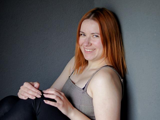 LaylaXDream, avec des cheveux roux, souriante, portant un débardeur rayé et un pantalon noir, assise contre un mur gris, les mains sur ses genoux, montrant subtilement ses seins..