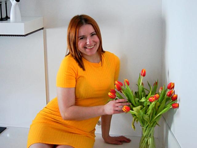 LaylaXDream con i capelli rossi, un vestito giallo, che sorride, tiene in mano tulipani arancioni e rossi, il braccio destro appoggiato su una superficie bianca, la mano sinistra sui tulipani, mostrando il suo sedere carino nella foto..