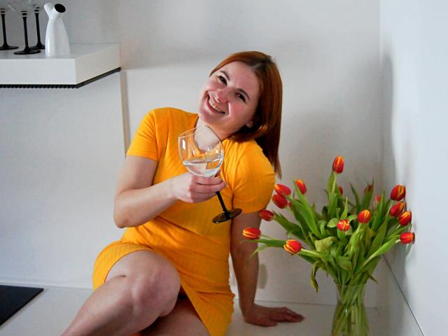 LaylaXDream con i capelli rossi, sorridente, che indossa un vestito giallo, tiene un bicchiere, seduta su un pavimento bianco, accanto a un vaso di tulipani arancioni e a un muro bianco..