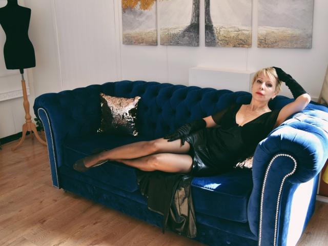 JustEva lounging on a blue velvet couch, wearing a black dress, sheer stockings, and black gloves, with her legs crossed, showing off her tits and ass.