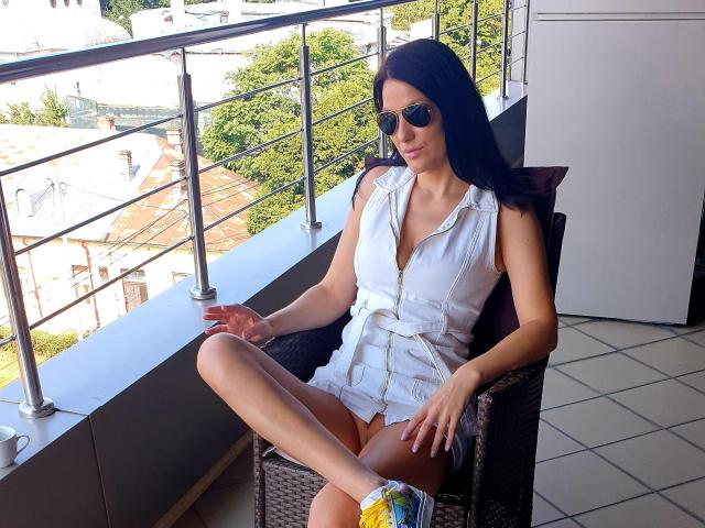 JoliDanielle with dark hair, sunglasses, white sleeveless dress, and yellow sneakers, sitting on a wicker chair, legs crossed, on a balcony with metal railing, and tiled floor.