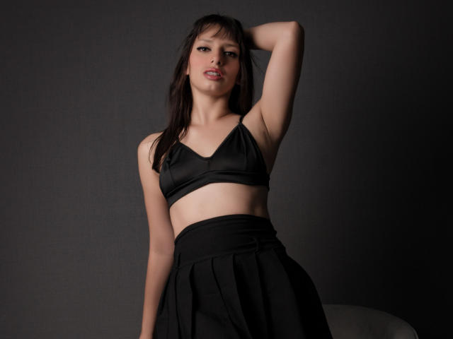 LeticiaFlorenz, con i capelli scuri, indossa un reggiseno e una gonna neri, con un braccio alzato che mostra il sedere scoperto, e la bocca leggermente aperta, su uno sfondo scuro..