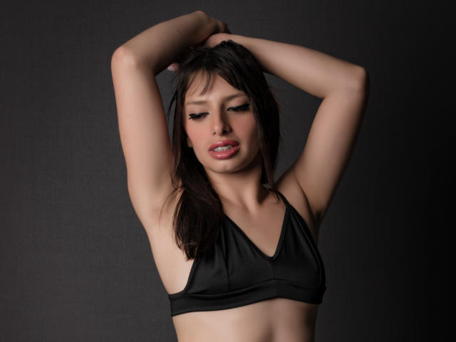 LeticiaFlorenz tem os braços levantados, vestindo um top de ginástica preto, expondo seus pequenos seios e mostrando seus lábios rosados, ligeiramente entreabertos, com uma expressão sutil e sexy..