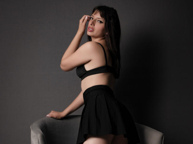 LeticiaFlorenz with dark hair, wearing a black bra and skirt, sits on a chair, hand on her face, looking over her shoulder, showing her ass and small tits.