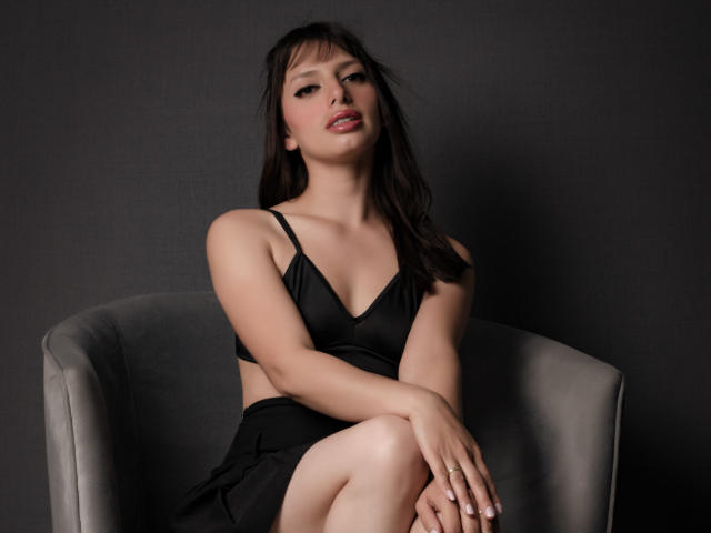 LeticiaFlorenz, com seios pequenos, vestindo um sutiã e saia pretos, sentada em uma cadeira cinza, com as mãos nos joelhos, fundo escuro, sorrindo levemente, cabelo castanho escuro, pele clara..
