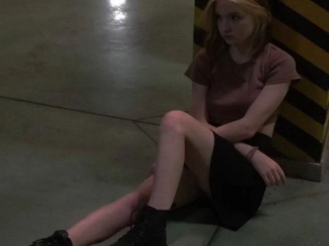 LaPochka, avec une peau pâle, des cheveux roux, portant un crop top marron, une jupe noire et des bottes noires, assise les jambes croisées, regardant légèrement sur le côté, dans une pièce faiblement éclairée..