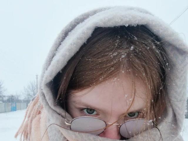 Das Gesicht von LaPochka mit Schnee in ihrem braunen Haar, sie trägt eine graue Jacke mit Kapuze, eine Brille, die ihre grünen Augen teilweise bedeckt, in einer verschneiten Outdoor-Umgebung..