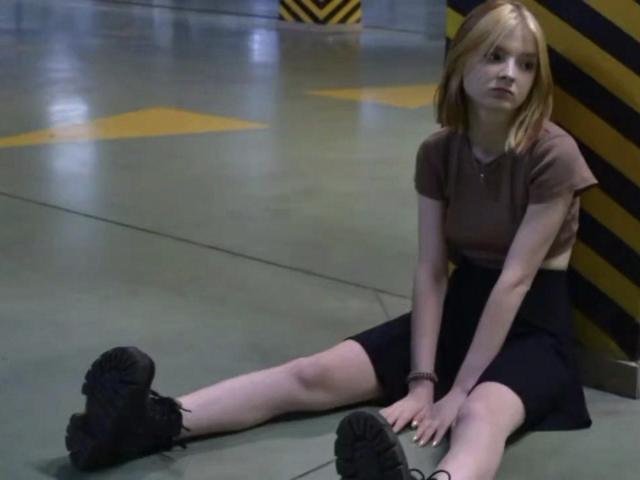 LaPochka mit blonden Haaren, einem braunen Hemd, einem schwarzen Rock und schwarzen Schuhen, sitzt auf dem Betonboden, die Beine gespreizt, und blickt nach oben, in einem schwach beleuchteten Industriezimmer..