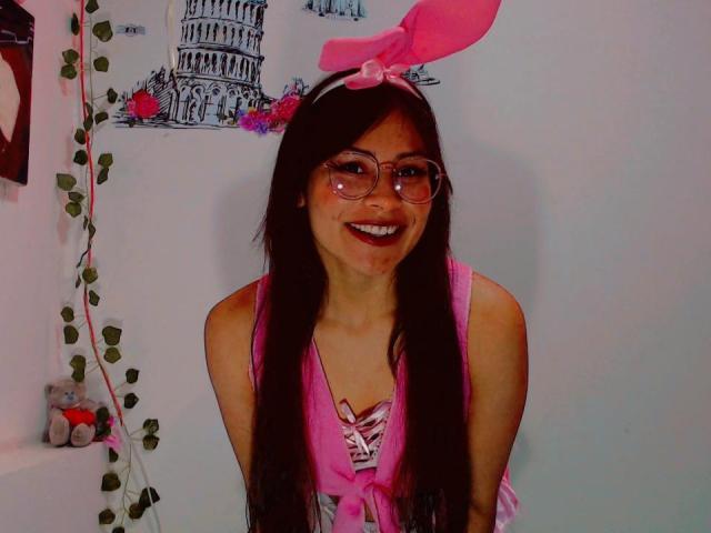 JadeFiore with long black hair, pink bow, and glasses, smiling, wearing a pink top, showing her tits, standing, pink nail polish, cute, sexy, pink-framed glasses, white wall.