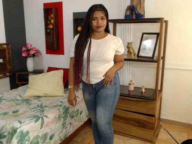 CataleyaX69, con largas trenzas oscuras, camisa blanca con textura, jeans azules, de pie en un dormitorio, con una mano en la cadera, mirando a la cámara, un pequeño reloj dorado, linda, con el trasero ligeramente visible..