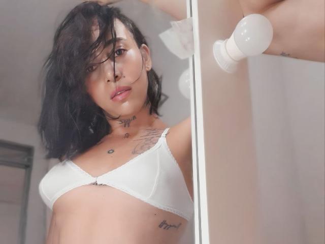 YeiSela with short black hair, white bra, small tits, tattoos on neck and chest, standing in a glass shower, looking at camera, light bulb in background.