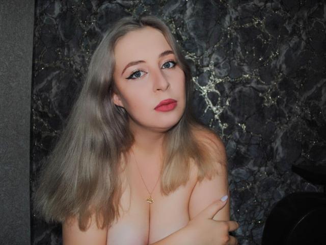 SweetLilit mit grauen Haaren, blauen Augen, rotem Lippenstift und einer goldenen Halskette, die ihre Brüste mit ihren Armen bedeckt und in die Kamera schaut. Marmorner Hintergrund..