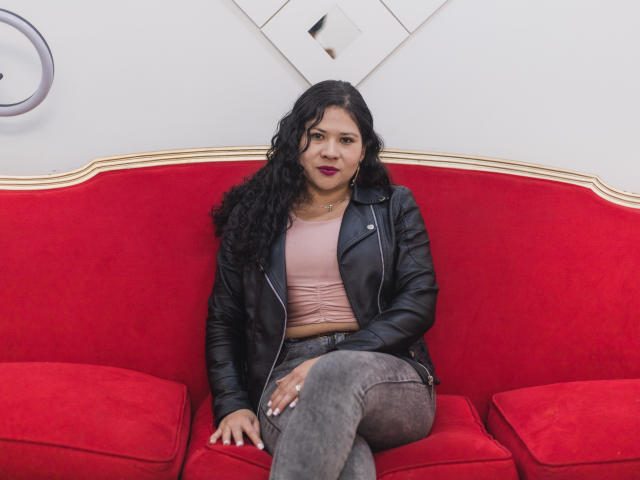 LourrieAnn, avec ses cheveux noirs bouclés, son haut rose, sa veste en cuir noir et son jean gris, assise sur un canapé rouge, dégage une ambiance confiante, séduisante et détendue..
