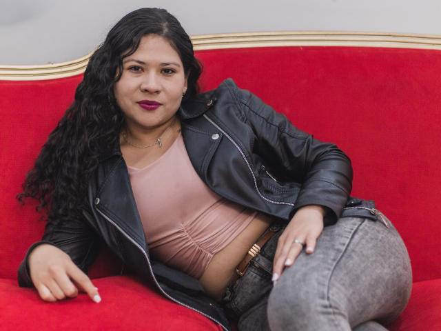 LourrieAnn mit lockigem, schwarzem Haar, die eine schwarze Lederjacke, ein pinkes Oberteil und graue Jeans trägt, sitzt entspannt auf einem roten Samtsessel, zeigt mit ihrer rechten Hand und trägt ein bezauberndes Lächeln..