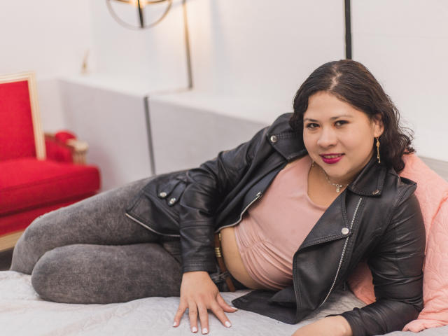 LourrieAnn, con una chaqueta de cuero negro, una blusa rosa, leggings grises y lápiz labial rojo, acostada en la cama, sonriendo, mostrando su suave trasero, sus pechos medianos y su cuerpo curvilíneo..