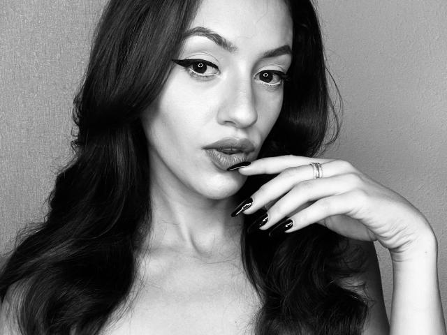 Vievienne Clemente, con cabello largo y oscuro, delineador de ojos alado y labios brillantes, tocándose los labios con dedos con uñas negras, llevando un anillo en el dedo índice, en una fotografía en blanco y negro..