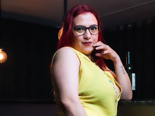 Sammy Owens, avec des cheveux roux, des lunettes et un haut jaune, montrant un tatouage à son poignet droit, touchant son menton, regardant la caméra, avec une bouteille en arrière-plan..