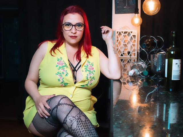 SammyOwens with red hair, glasses, yellow dress, black lace bra, fishnet stockings, sitting on counter, hand on knee, bottle and glasses on counter, smiling, tits visible.