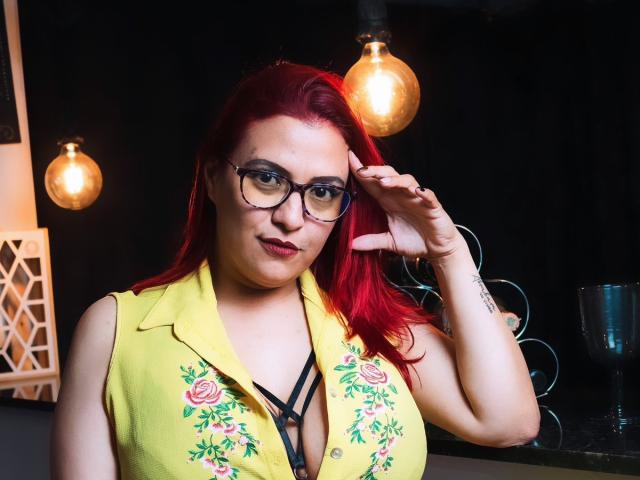 SammyOwens with red hair, glasses, yellow floral vest, black strappy top, big boobs, fair skin, touching hair, black background, hanging lights, tattoo on wrist, confident smile.