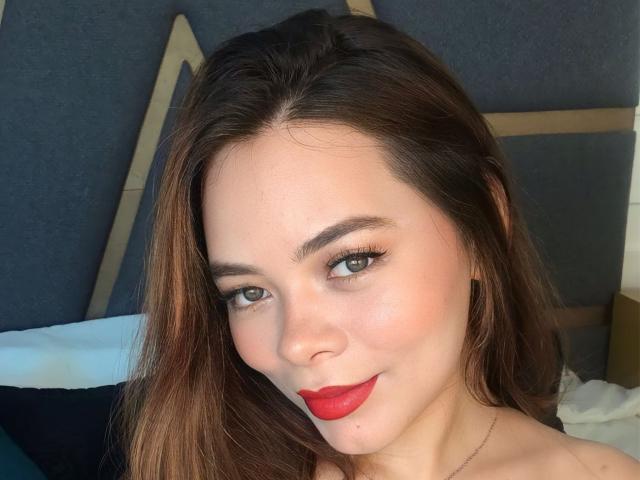 Anika Gilbert tiene piel clara, ojos verdes y lápiz labial rojo. Está sonriendo, con el pelo largo y castaño y un maquillaje discreto. Sus senos no son visibles, solo su rostro..