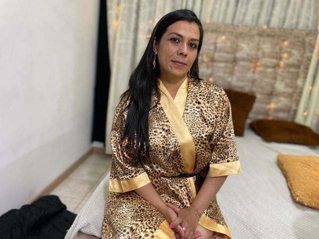 MartinaFox69, donna dai capelli scuri, indossa un vestaglie a stampa leopardo con bordi dorati, seduta sul letto, mani sulle ginocchia, occhi marroni, leggero sorriso, orecchini dorati, capelli neri sciolti, ambientazione: camera da letto..