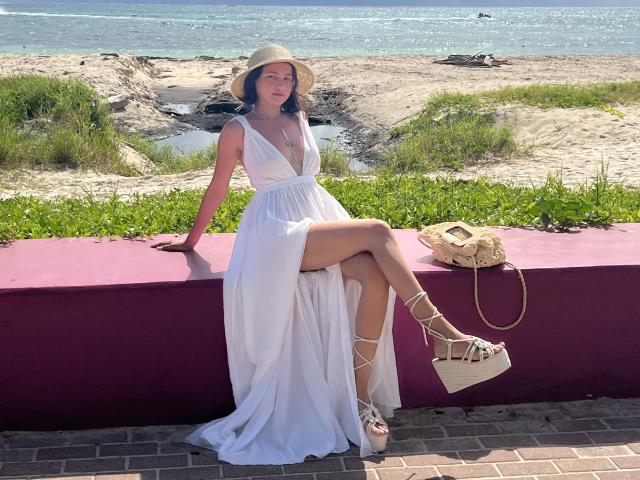 EstefaniaRamosHot sits on a pink wall in a white dress, showing her tits in a deep V, with her leg crossed, wearing wedge sandals, and a sunhat.