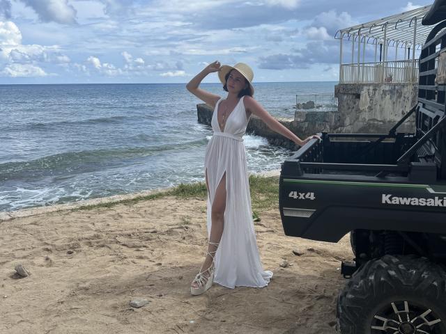 EstefaniaRamosHot è in piedi, indossa un abito bianco con un profondo scollo e uno spacco laterale, porta un cappello che tiene in mano e posa accanto a un quad Kawasaki verde su una spiaggia sabbiosa..