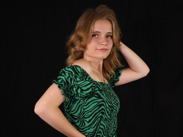 MargaritaKis with wavy blonde hair, green zebra-print dress, hand in hair, confident smirk, black background. Boobs slightly visible under dress. She's cute, has light skin.